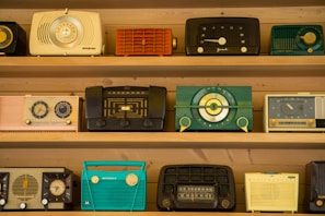 A collection of vintage radios is displayed on wooden shelves. Each radio has a distinct design and color, ranging from teal and black to yellow and red. The styles reflect a bygone era with dials, knobs, and mesh speaker coverings, showcasing the aesthetic diversity of mid-20th-century radio craftsmanship.