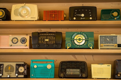 A collection of vintage radios is displayed on wooden shelves. Each radio has a distinct design and color, ranging from teal and black to yellow and red. The styles reflect a bygone era with dials, knobs, and mesh speaker coverings, showcasing the aesthetic diversity of mid-20th-century radio craftsmanship.