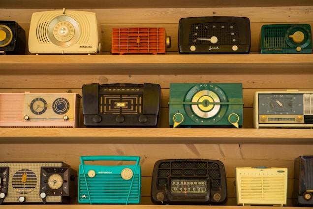 A collection of vintage radios is displayed on wooden shelves. Each radio has a distinct design and color, ranging from teal and black to yellow and red. The styles reflect a bygone era with dials, knobs, and mesh speaker coverings, showcasing the aesthetic diversity of mid-20th-century radio craftsmanship.
