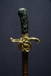 A decorative sword with an intricately carved handle and a gold-colored guard. The handle features detailed engravings, possibly depicting scenes or figures. The gold guard is ornate, adding a luxurious touch to the weapon.