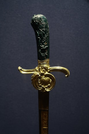 A decorative sword with an intricately carved handle and a gold-colored guard. The handle features detailed engravings, possibly depicting scenes or figures. The gold guard is ornate, adding a luxurious touch to the weapon.