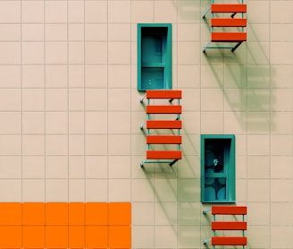 A building facade with a grid of square panels in beige, featuring rectangular windows with blue-green frames. Three orange-red external staircases or platforms are attached below the windows, casting shadows on the wall. The bottom row of panels includes a contrasting orange section.