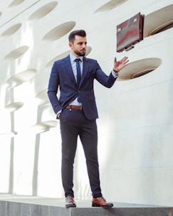A man in a dark blue suit is standing against a modern architectural wall with oval cutouts. He is balancing a brown briefcase in mid-air with one hand while his other hand is in his pocket. His attire includes a matching tie, a white shirt, and brown leather shoes. The lighting creates soft shadows on the wall, enhancing the contemporary feel.