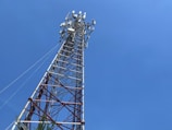 A tall telecommunications tower extends upward against a clear blue sky. The tower is composed of metal latticework with multiple crossbars and various antennas attached near the top. This structure is anchored by cables providing stability. At the base, there is some greenery visible.