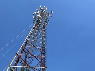 A tall telecommunications tower extends upward against a clear blue sky. The tower is composed of metal latticework with multiple crossbars and various antennas attached near the top. This structure is anchored by cables providing stability. At the base, there is some greenery visible.