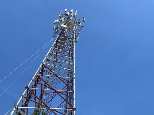A tall telecommunications tower extends upward against a clear blue sky. The tower is composed of metal latticework with multiple crossbars and various antennas attached near the top. This structure is anchored by cables providing stability. At the base, there is some greenery visible.