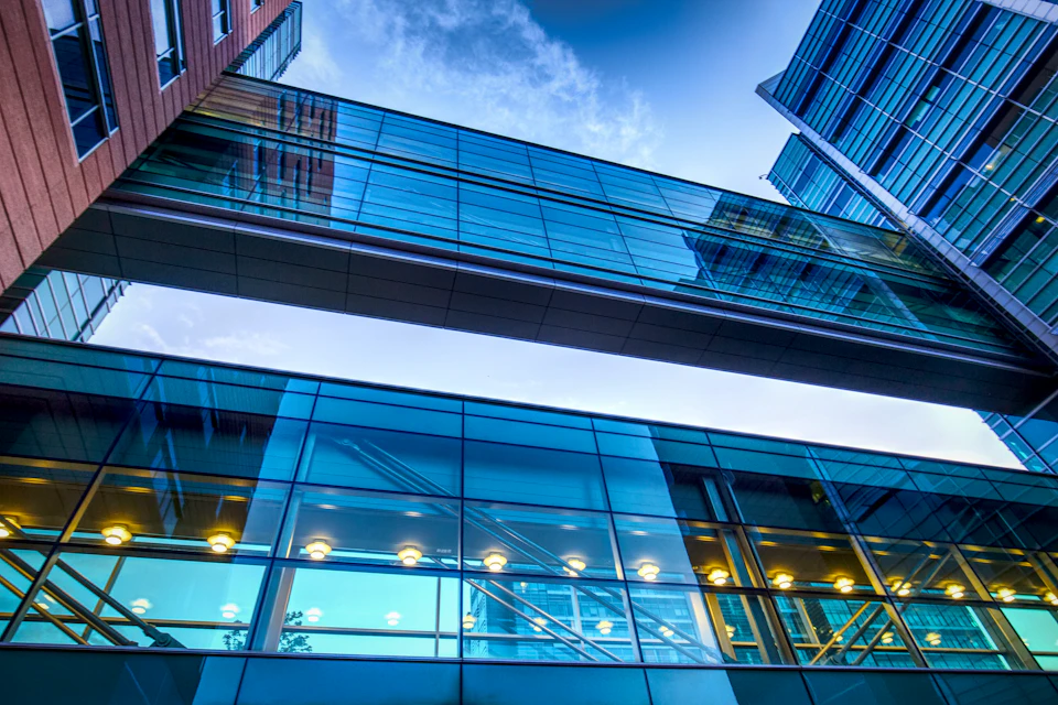 Modern glass and steel building with walkway connecting two levels under evening illumination