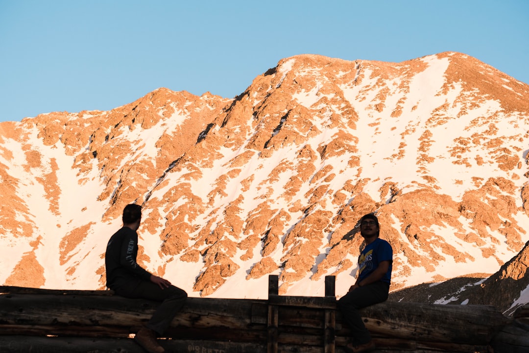 man in black jacket sitting on brown wooden fence looking at brown and white mountain during