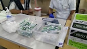 A table with medical supplies including cotton balls, disposable syringes, and hand sanitizer. Two individuals are partially visible, likely medical staff, one wearing a white coat and gloves. Cardboard boxes are stacked on the side.