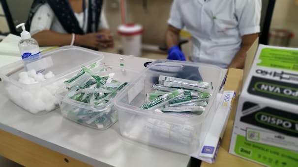 A table with medical supplies including cotton balls, disposable syringes, and hand sanitizer. Two individuals are partially visible, likely medical staff, one wearing a white coat and gloves. Cardboard boxes are stacked on the side.