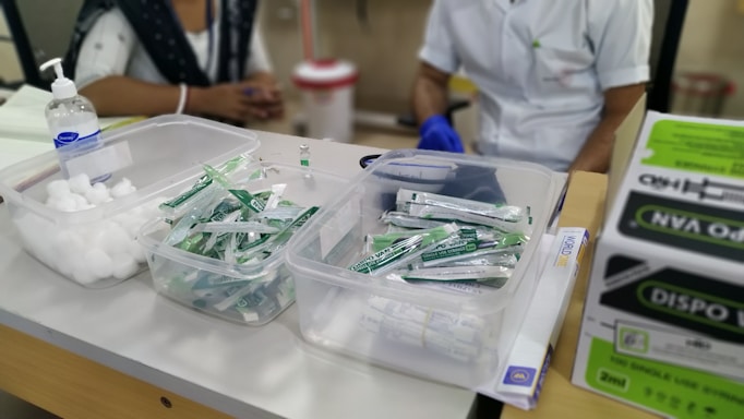 A table with medical supplies including cotton balls, disposable syringes, and hand sanitizer. Two individuals are partially visible, likely medical staff, one wearing a white coat and gloves. Cardboard boxes are stacked on the side.