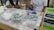 A table with medical supplies including cotton balls, disposable syringes, and hand sanitizer. Two individuals are partially visible, likely medical staff, one wearing a white coat and gloves. Cardboard boxes are stacked on the side.