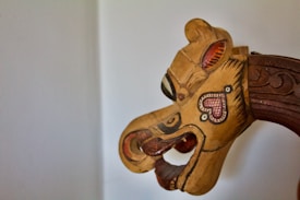 A wooden carved puppet head with intricate painted details. The design features bold, contrasting colors and a stylized heart symbol on the cheek. The features include prominent eyes and a mouth with an open, expressive shape. The craftsmanship is intricate with distinct patterns and shapes.