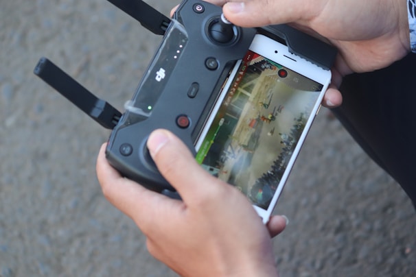 Close-up of hands holding a remote controller for a drone or similar device. The controller is connected to a smartphone displaying a live video feed or screen of a game. The background is out of focus.