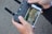 Close-up of hands holding a remote controller for a drone or similar device. The controller is connected to a smartphone displaying a live video feed or screen of a game. The background is out of focus.