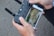 Close-up of hands holding a remote controller for a drone or similar device. The controller is connected to a smartphone displaying a live video feed or screen of a game. The background is out of focus.