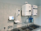 A laboratory setup featuring a water purification system mounted on a tiled wall above a stainless steel double sink. The setup includes two square white units labeled with blue text, and interconnected pipes and blue water filters beneath. A touch screen control panel is installed on the left side with a digital display.