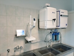 A laboratory setup featuring a water purification system mounted on a tiled wall above a stainless steel double sink. The setup includes two square white units labeled with blue text, and interconnected pipes and blue water filters beneath. A touch screen control panel is installed on the left side with a digital display.