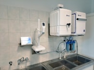 A laboratory setup featuring a water purification system mounted on a tiled wall above a stainless steel double sink. The setup includes two square white units labeled with blue text, and interconnected pipes and blue water filters beneath. A touch screen control panel is installed on the left side with a digital display.