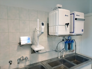 A laboratory setup featuring a water purification system mounted on a tiled wall above a stainless steel double sink. The setup includes two square white units labeled with blue text, and interconnected pipes and blue water filters beneath. A touch screen control panel is installed on the left side with a digital display.