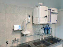 A laboratory setup featuring a water purification system mounted on a tiled wall above a stainless steel double sink. The setup includes two square white units labeled with blue text, and interconnected pipes and blue water filters beneath. A touch screen control panel is installed on the left side with a digital display.