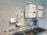 A laboratory setup featuring a water purification system mounted on a tiled wall above a stainless steel double sink. The setup includes two square white units labeled with blue text, and interconnected pipes and blue water filters beneath. A touch screen control panel is installed on the left side with a digital display.