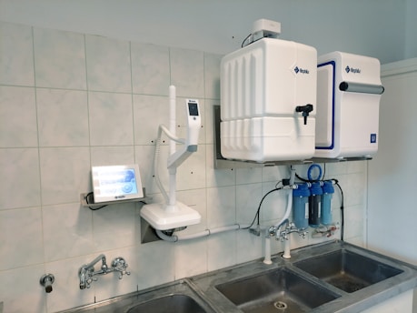 A laboratory setup featuring a water purification system mounted on a tiled wall above a stainless steel double sink. The setup includes two square white units labeled with blue text, and interconnected pipes and blue water filters beneath. A touch screen control panel is installed on the left side with a digital display.