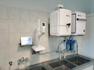 A laboratory setup featuring a water purification system mounted on a tiled wall above a stainless steel double sink. The setup includes two square white units labeled with blue text, and interconnected pipes and blue water filters beneath. A touch screen control panel is installed on the left side with a digital display.