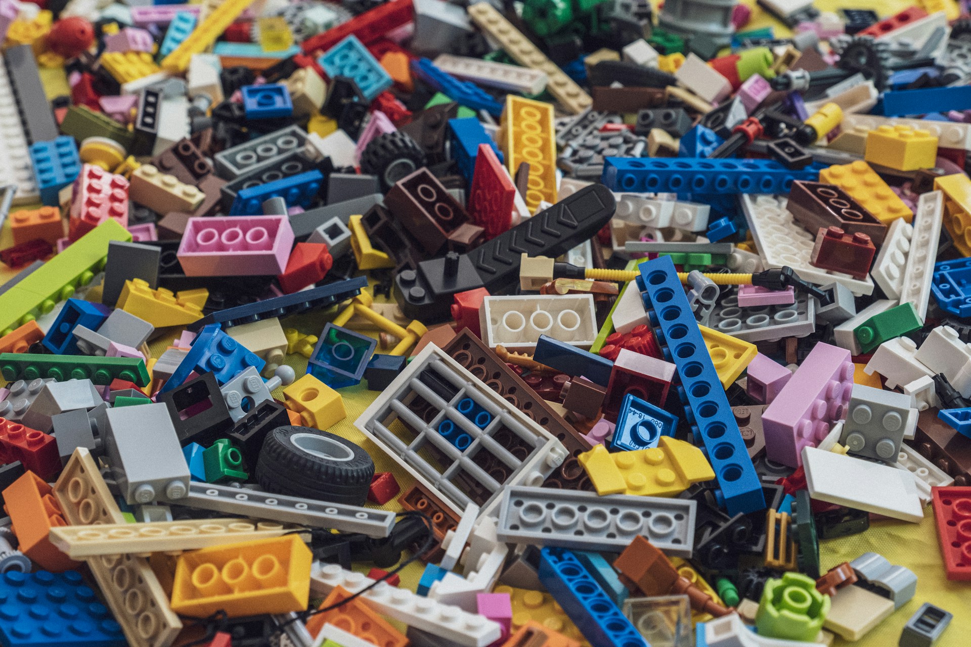 A large pile of multicolored LEGO bricks and pieces is scattered across a surface. The bricks are of various shapes, sizes, and colors, including reds, blues, greens, yellows, and more. Some pieces are recognizable as wheels, windows, and other specific LEGO elements.