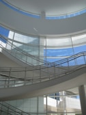 A modern architectural interior with a sleek, curving staircase and large windows allowing abundant natural light. The structure features a minimalist design with an emphasis on clean lines and open space.