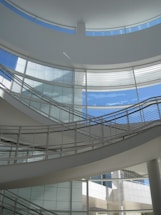 A modern architectural interior with a sleek, curving staircase and large windows allowing abundant natural light. The structure features a minimalist design with an emphasis on clean lines and open space.