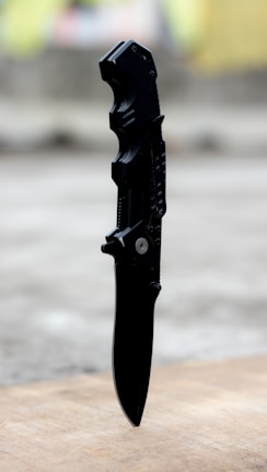 A rugged survival knife resting on a wooden surface with outdoor gear in the background.