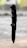 A black folding knife with a tactical tanto blade is positioned vertically on a wooden surface. The knife features a textured handle and appears sharp and sturdy. The background is blurred, suggesting an outdoor setting.