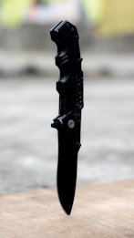 A black folding knife with a tactical tanto blade is positioned vertically on a wooden surface. The knife features a textured handle and appears sharp and sturdy. The background is blurred, suggesting an outdoor setting.