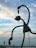 A large abstract sculpture with elongated arms holds a fish in each hand, set against a backdrop of a partly cloudy sky. In the distance, a crane and part of a building or pier are visible.