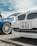 A close-up view of the front ends of two cars parked side by side. The focus is on a white car with a Japanese license plate and an Alpina badge. The blue car next to it has a sleek, stylish design with large alloy wheels. The background shows a cloudy sky and some greenery.