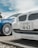 A close-up view of the front ends of two cars parked side by side. The focus is on a white car with a Japanese license plate and an Alpina badge. The blue car next to it has a sleek, stylish design with large alloy wheels. The background shows a cloudy sky and some greenery.