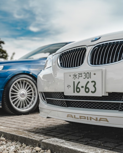 A close-up view of the front ends of two cars parked side by side. The focus is on a white car with a Japanese license plate and an Alpina badge. The blue car next to it has a sleek, stylish design with large alloy wheels. The background shows a cloudy sky and some greenery.