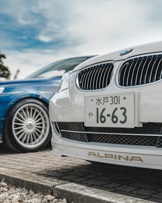 A close-up view of the front ends of two cars parked side by side. The focus is on a white car with a Japanese license plate and an Alpina badge. The blue car next to it has a sleek, stylish design with large alloy wheels. The background shows a cloudy sky and some greenery.