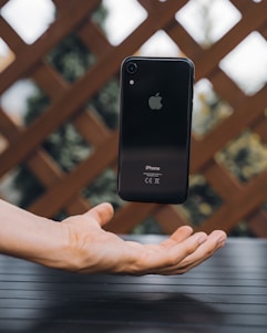 A black iPhone is suspended in mid-air above an outstretched hand, creating the illusion of levitation. The background consists of a patterned wooden fence and out-of-focus greenery.