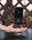 A black iPhone is suspended in mid-air above an outstretched hand, creating the illusion of levitation. The background consists of a patterned wooden fence and out-of-focus greenery.