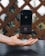 A black iPhone is suspended in mid-air above an outstretched hand, creating the illusion of levitation. The background consists of a patterned wooden fence and out-of-focus greenery.