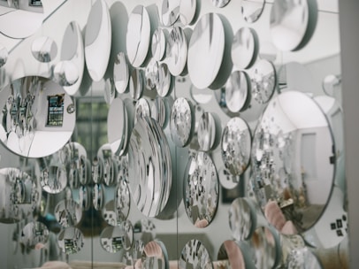 Numerous circular mirrors of varying sizes are mounted on a wall, creating an artistic and visually striking pattern of reflections. The mirrors reflect parts of the surrounding room, introducing a fragmented and abstract image of the environment, including furniture and decor.