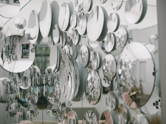 Numerous circular mirrors of varying sizes are mounted on a wall, creating an artistic and visually striking pattern of reflections. The mirrors reflect parts of the surrounding room, introducing a fragmented and abstract image of the environment, including furniture and decor.