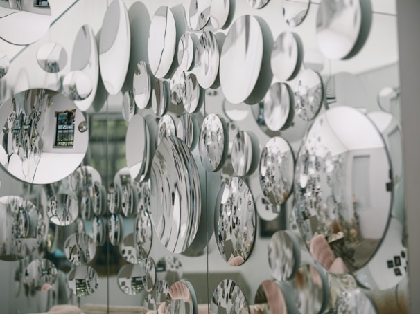 Numerous circular mirrors of varying sizes are mounted on a wall, creating an artistic and visually striking pattern of reflections. The mirrors reflect parts of the surrounding room, introducing a fragmented and abstract image of the environment, including furniture and decor.