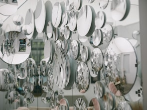 Numerous circular mirrors of varying sizes are mounted on a wall, creating an artistic and visually striking pattern of reflections. The mirrors reflect parts of the surrounding room, introducing a fragmented and abstract image of the environment, including furniture and decor.