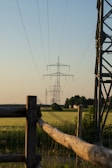 A series of electrical transmission towers extend into the distance over a flat, green field. Wooden fences and metal structures are also present on the sides of the image. The sky is clear with a soft gradient from blue to a warmer hue near the horizon, indicating a possible early morning or late afternoon setting.