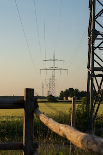 A series of electrical transmission towers extend into the distance over a flat, green field. Wooden fences and metal structures are also present on the sides of the image. The sky is clear with a soft gradient from blue to a warmer hue near the horizon, indicating a possible early morning or late afternoon setting.