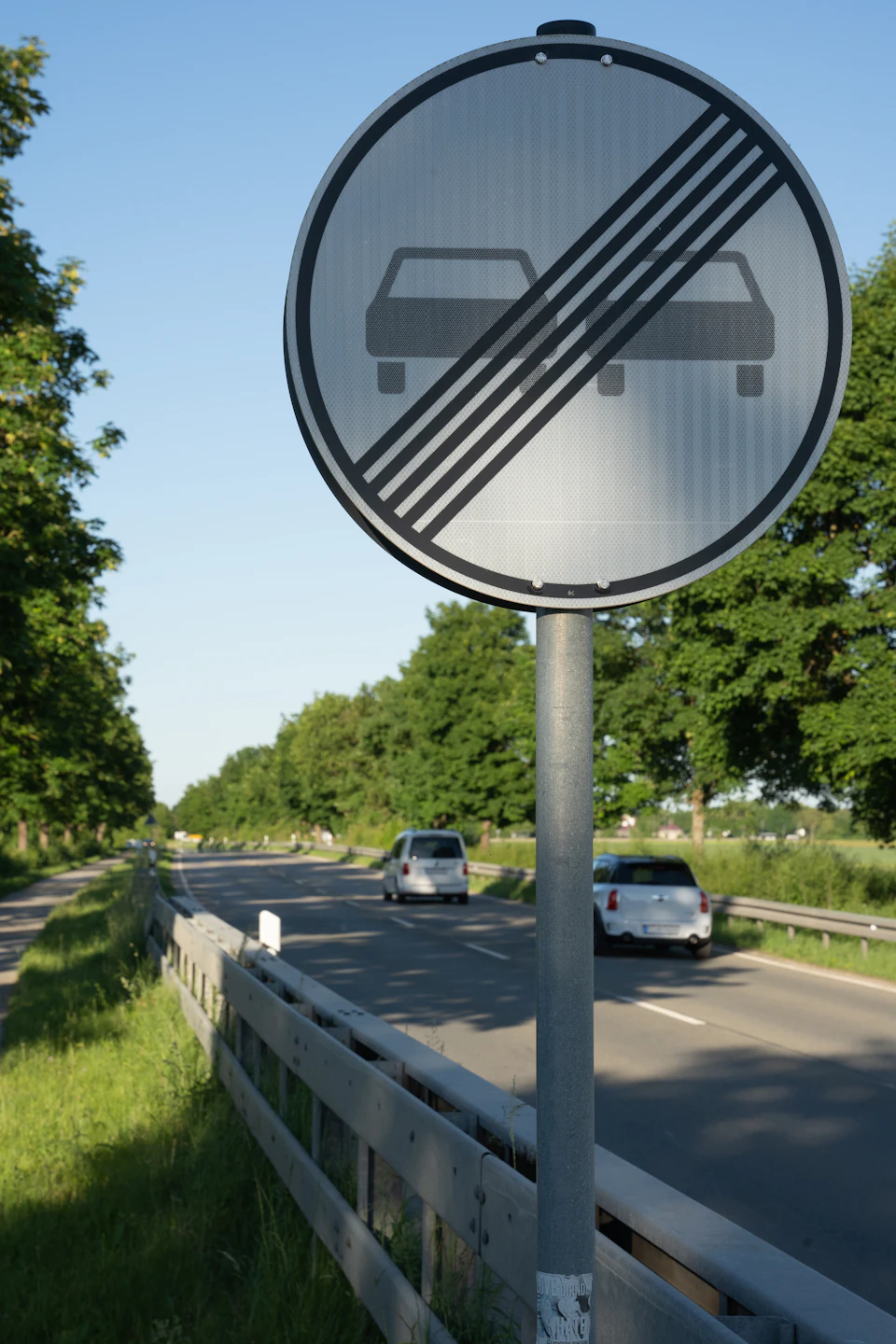 The image shows a two-lane road with a "No Parking" sign on the right side, indicating that parking is not allowed in that area. The road is surrounded by lush green trees and grass, creating a serene atmosphere. On the left side of the road, there are three cars parked along the curb, while on the right side, two more cars can be seen further down the road.