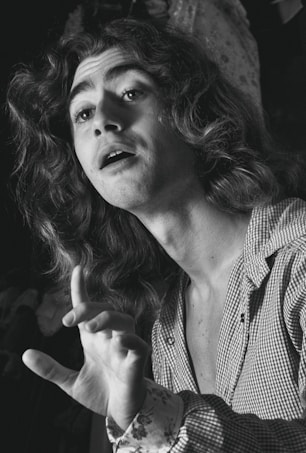 A black and white image of a person with long wavy hair, wearing a checked shirt, and gesturing with one hand. The person appears to be in a dramatic pose, possibly singing or speaking.